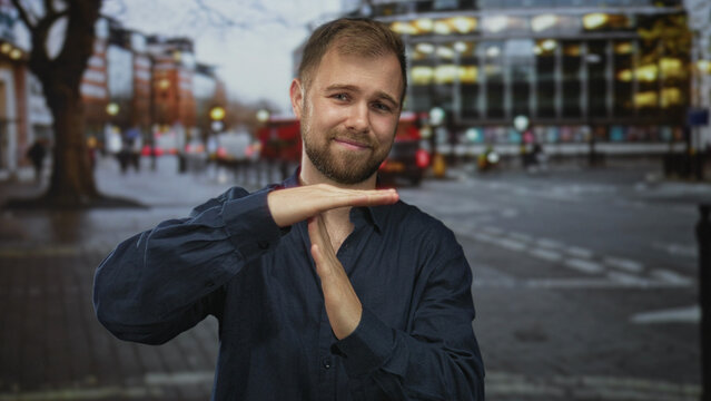 Man with beard making time out gesture with both hands and slight smile, wearing navy shirt, standing on busy city street at dusk; playful pause.