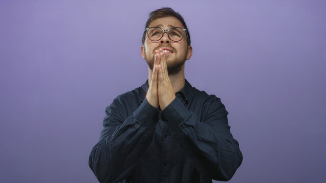 Man wearing glasses with hands pressed in prayer in purple studio looking upward; hope prayer plea sincere.