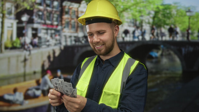 Man builder in hardhat counting dollar bills with hands on street by canal and bridge in amsterdam; pride financial success.