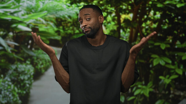 Man in black t shirt shrugs with palms up on a leafy green park path, facing camera with a slight smile; uncertainty casual relaxed.