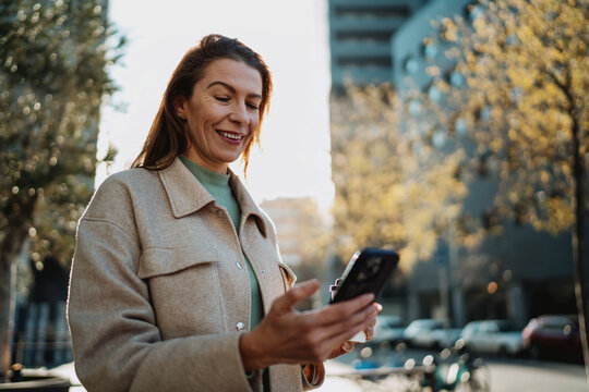 Happy lady looking at smartphone screen holding cup of coffee outdoors. Modern woman using mobile internet for social network communication or shopping online. Authentic professional lifestyle.