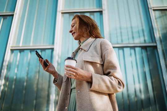 Low angle view mature woman using smartphone app holding paper coffee cup. Focused female entrepreneur browsing mobile phone screen near modern glass facade office building. Digital nomad life.