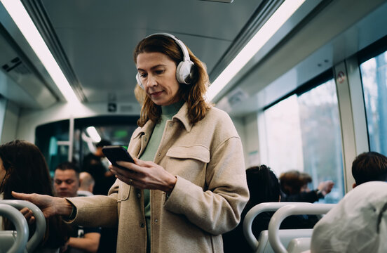 Caucasian middle aged woman standing inside tram, using smartphone app, wearing white headphones, commuting in public transport, holding handle, digital lifestyle, looking at phone screen.