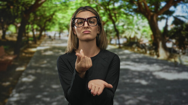 Woman in black sweater points thumbs to chest in sunlit park path while wearing oversized glasses and puzzled expression; self doubt confusion.
