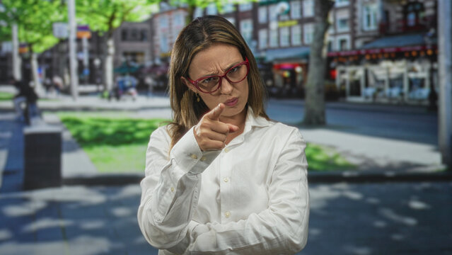 Woman in white shirt and red glasses points finger forward with crossed arms on city street with buildings; accusation doubt confrontation.