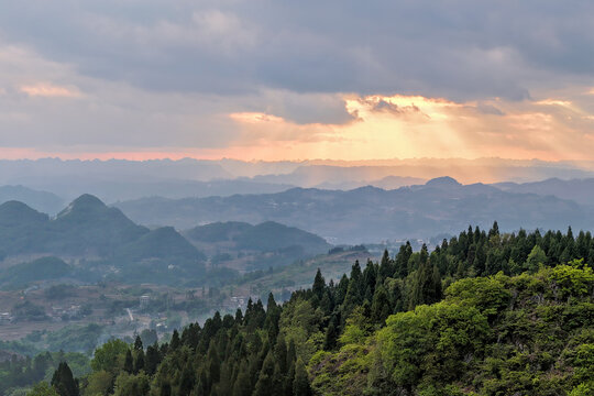 Aerial view of rolling hills and dense evergreen forests under a dramatic sky with sunbeams breaking through the clouds over Gaoxing Village, Guizhou, China.
