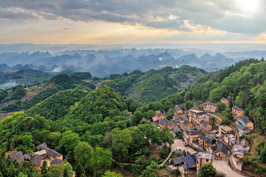 Aerial view of traditional houses in a remote mountain village surrounded by lush green karst peaks and misty valleys under a cloudy sky in Gaoxing Village, Guizhou, China.