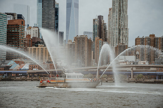 FDNY fireboat water cannon display in New York City harbor. Close view of rescue vessel spraying high pressure water jets with Manhattan skyline and skyscrapers behind.