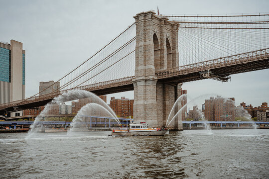 FDNY fireboat spraying water cannons near Brooklyn Bridge New York. Marine fireboat performing water display on East River under suspension bridge in New York City.