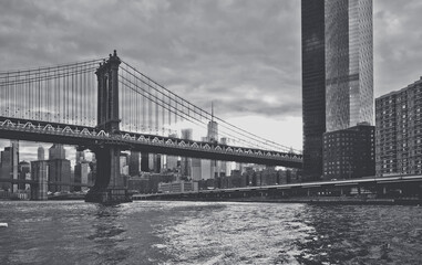 Naklejka premium Black and white view of Manhattan Bridge and skyline. Monochrome image of suspension bridge, skyscrapers, and river waves, high contrast, cloudy sky, urban scene.