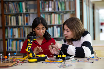 Fototapeta premium Indian girls focus intently on robotic arm project. hands adjusting wires and parts. The library setting suggests learning or innovation. Their expressions show curiosity. Education, Back to school