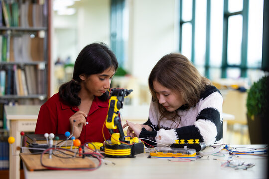 Indian girls focus intently on robotic arm project. hands adjusting wires and parts. The library setting suggests learning or innovation. Their expressions show curiosity. Education, Back to school