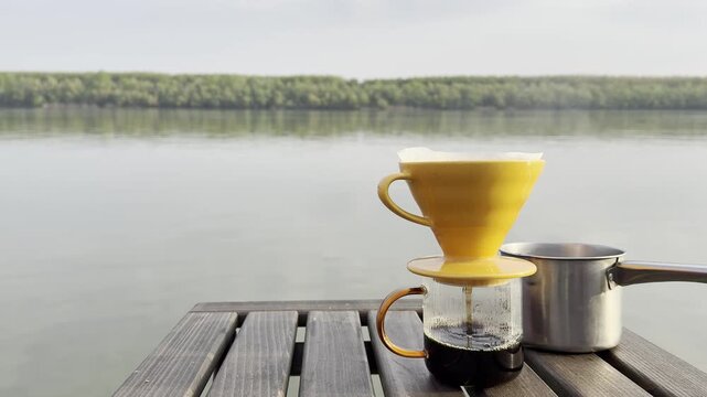 Close-up of hand pouring hot water into a pour over coffee dripper on a wooden table near a peaceful lake. Minimal outdoor coffee brewing scene with calm water, forest landscape, and soft natural