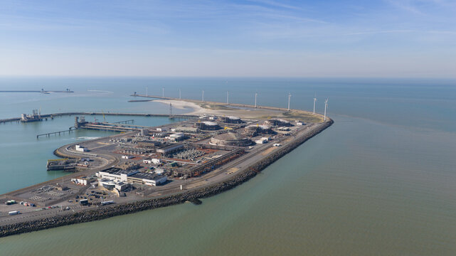 Aerial view of the Fluxys LNG Terminal Zeebrugge with large storage tanks and wind turbines along the coastline under a clear blue sky in Brugge, Vlaanderen, Belgium.