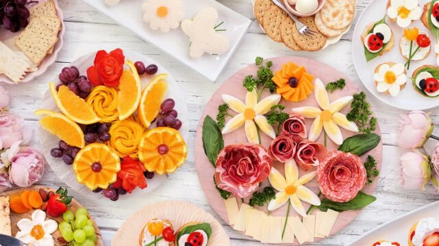 Summer garden party food table scene. Slow rotating zoom motion. Variety of fun flower theme charcuterie items, fruit, crostini, finger sandwiches and dessert. Top down view on a white wood background
