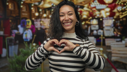 Woman forming heart with hands on a busy street market stall, wearing striped sweater and hoop earrings, smiling warmly  love joy. © Krakenimages.com
