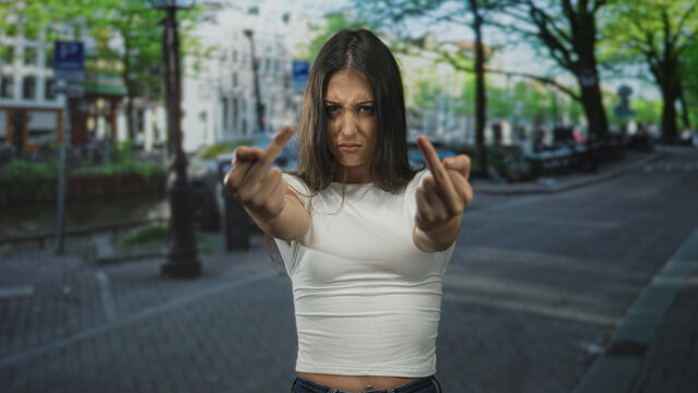 Woman holding up both middle fingers toward camera on a city street with visible bellybutton piercing and tense facial expression; defiance.