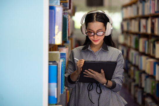 Young Asian woman smiles while listening to music in library. She wears glasses and headphones, Bookshelves line both sides of the aisle behind her. Soft light illuminates her focused. Education