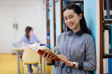 Fototapeta premium Young Asian woman studies in quiet library. Books line tall shelves behind. she holds book, eyes focused. Serene atmosphere, calm concentration. Ideal for educational or study-themed projects