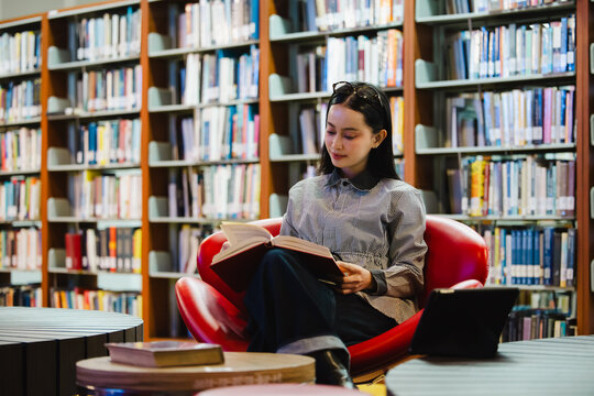 Young Asian woman studies reading book anf Siting on red chair in quiet library. Books line tall shelves behind. she holds book, calm concentration. Ideal for educational or study-themed projects