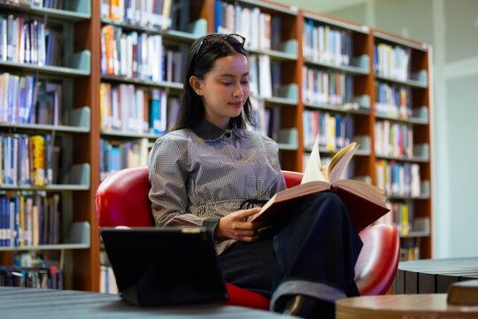 Young Asian woman studies reading book anf Siting on red chair in quiet library. Books line tall shelves behind. she holds book, calm concentration. Ideal for educational or study-themed projects