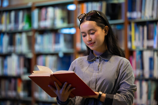 Young Asian woman studies reading book anf Siting on red chair in quiet library. Books line tall shelves behind. she holds book, calm concentration. Ideal for educational or study-themed projects