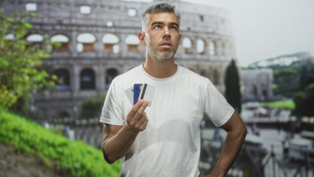 Man holding creditcard and debitcard in hand at historic building colosseum, facing camera with pensive look; financial anxiety.