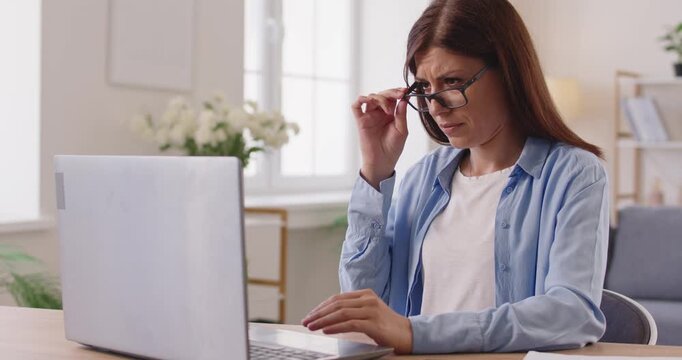 Woman with poor vision problem looking at laptop screen, sitting at desk at home. Worried person moving glasses to struggle with myopia, eye fatigue or presbyopia at computer, adjust focus for reading
