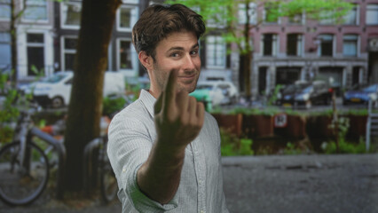 Fototapeta premium Young hispanic man shows middle finger to camera on a city street with bicycles and canal; defiance attitude.