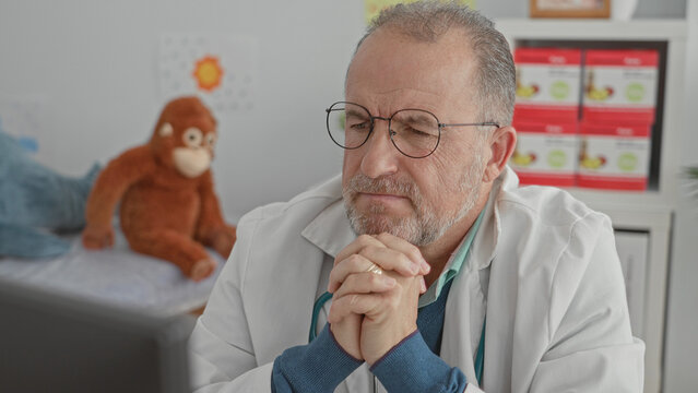 Man doctor with stethoscope, hands clasped and eyes closed at desk in clinic building with toy monkey and monitor visible; contemplation.