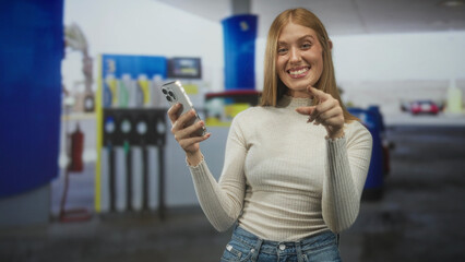 Young redhead woman smiling, holding smartphone, pointing finger at camera in gas station forecourt wearing light sweater and jeans  playful confidence. © Krakenimages.com