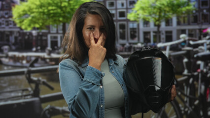 Fototapeta premium Woman holding motorbike helmet with two fingers to nose and pointing finger while standing on street by canal and parked bicycles; confidence independence.