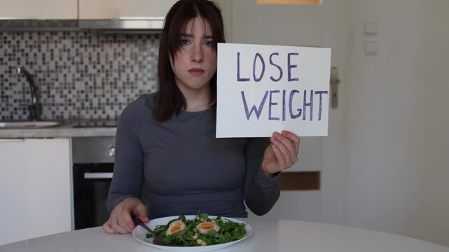 Young woman eats healthy salad while holding lose weight sign showing emotional pressure of modern beauty standards and toxic diet culture obsession