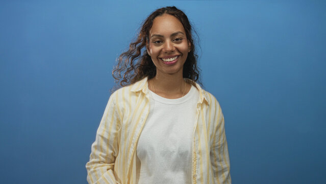 Woman with hand covering eyes and hand over mouth wearing white t shirt and yellow striped shirt in studio blue backdrop, mid gesture portrait; playful candid.