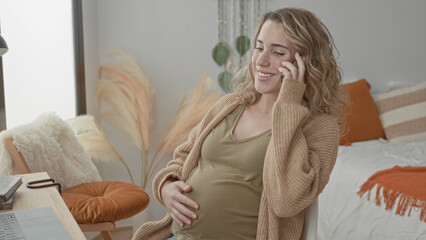 Pregnant woman talking on smartphone, smiling and cradling belly while seated at desk beside bed...