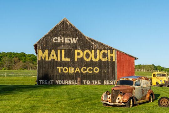 Iconic Mail Pouch tobacco barn featuring weathered wood and bold signage, with rusted vintage cars in front, surrounded by lush green grass under blue spring skies in Perkiomenville, PA.