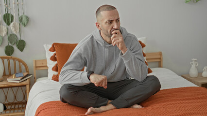 Fototapeta premium Man holds wrist and checks watch while seated crosslegged on bed beside nightstand and plant in a studio; concern.