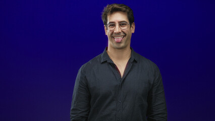 Obraz premium Man smiling with tongue out, face visible and wearing glasses with open collar shirt, slight head tilt, standing against blue studio backdrop; playful warmth.