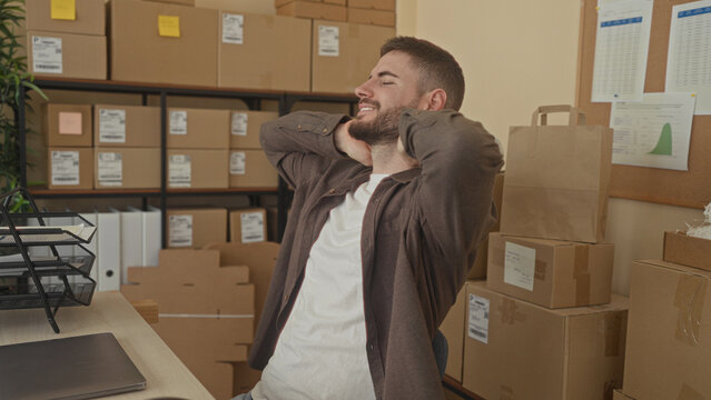 Man leans back with hands behind neck beside laptop and stacked packages in a small building office; relief small business.