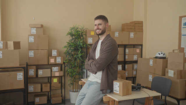 Man with arms crossed leaning on a parcel at a packing table surrounded by stacked cardboard boxes and shelving in a building; small business confidence.