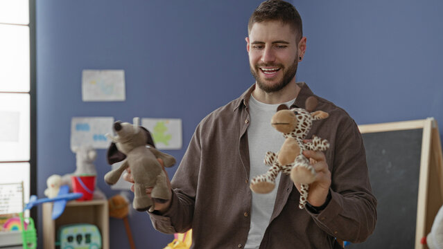 Young man holds two stuffed toys and puppets while smiling with open palms in a classroom building among shelves and a chalkboard; playful warmth.
