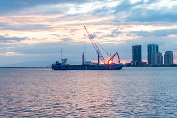 Fototapeta premium Construction barge with crane operates at sunset near modern city skyline