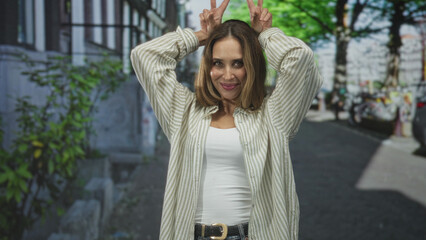 Fototapeta premium Woman smiling with hands making bunny ears above head on street wearing white tanktop and striped overshirt; playful urban moment.