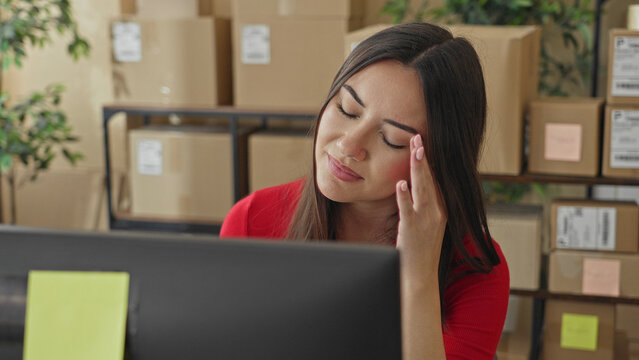 Woman rubbing temple with hand at computer among stacked parcels and boxes in building; small business fatigue.