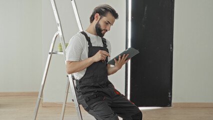 Man with beard and overalls points finger at tablet while seated on ladder inside a building near a black door panel and wooden floor  concentration. © Krakenimages.com