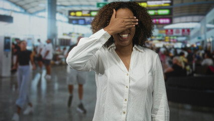 Fototapeta premium Woman covering eyes with hand while smiling in a busy airport terminal with check in kiosks and travelers behind her; lighthearted travel.