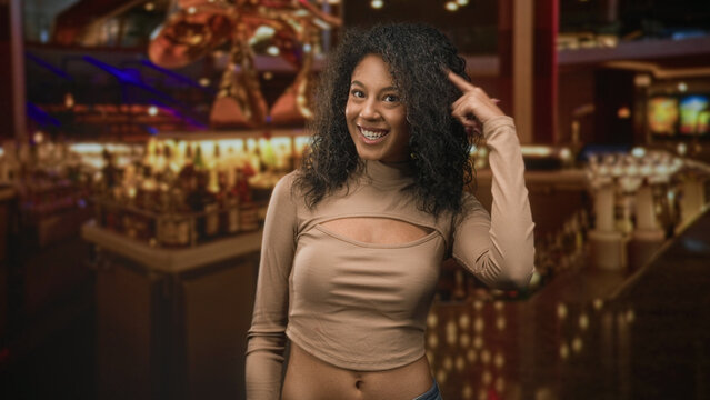Teenage woman shows midriff and points finger to head in casino building, smiling and animated; playful confidence.