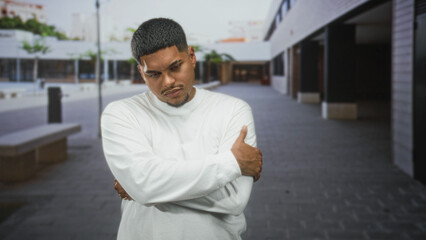 Fototapeta premium Hispanic man in white long sleeve shirt grips his arm, grimacing and holding elbow near concrete benches outside a commercial building plaza; discomfort.
