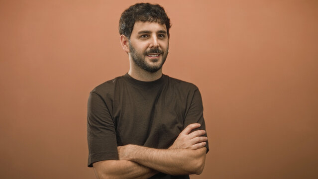 Man with beard crossing arms and slight smile in studio portrait with warm terracotta wall and dark tshirt; quiet confidence.