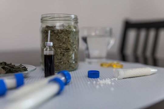 Close-up of homeopathic remedies on table: small glass vial with herbal tincture, open tube with homeopathic pellets, jar of dried herbs and glass of water. Natural medicine and holistic wellness.
